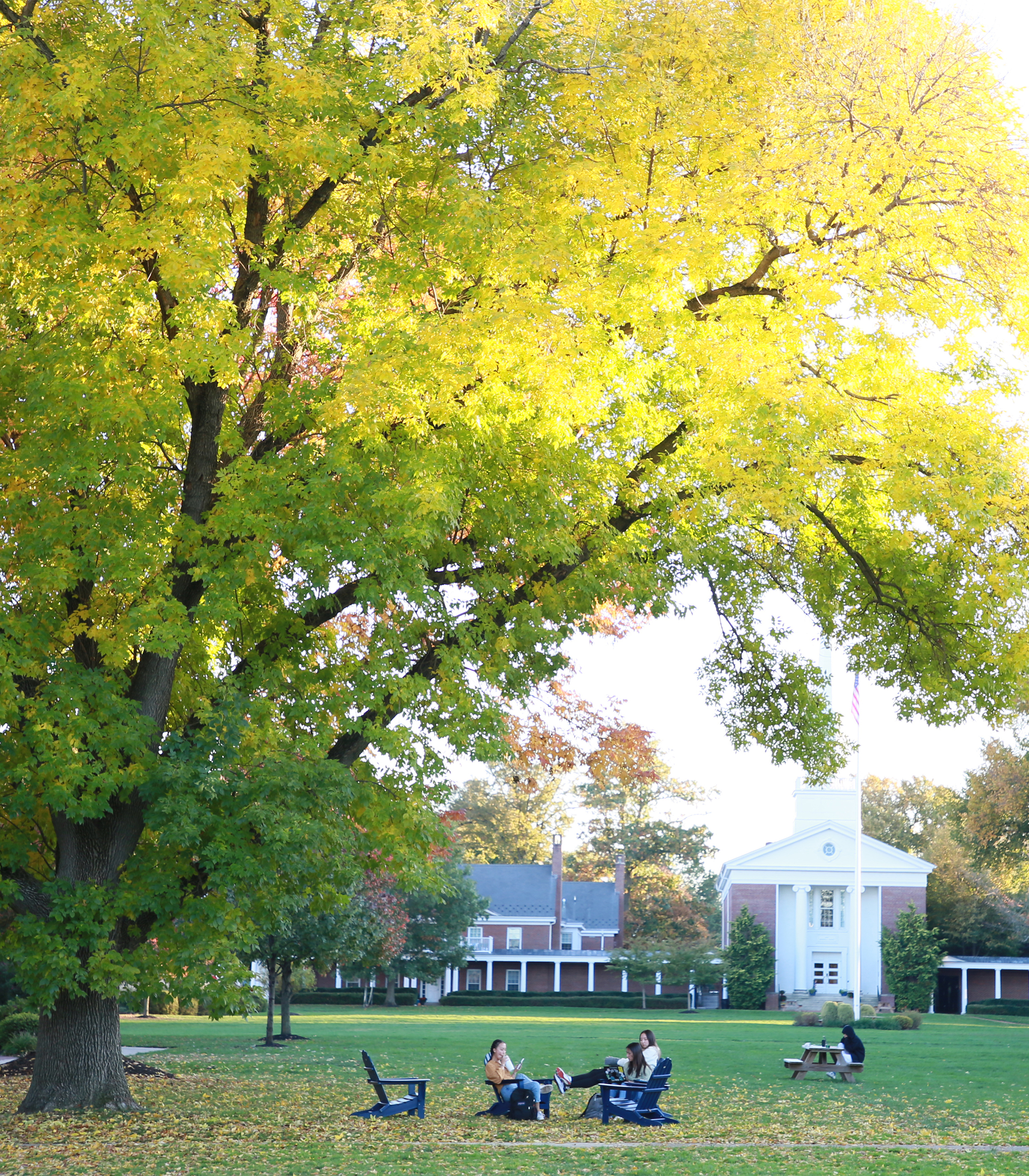 sunlight peering in through the green full trees with chapel framed in the background and students sitting on chairs in the foreground